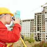 Land surveyor standing in front of a civil engineering building project, looking through a theodolite taking measurements.