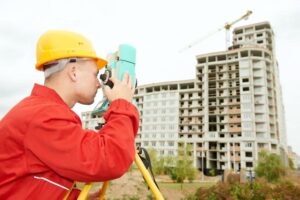 Land surveyor standing in front of a civil engineering building project, looking through a theodolite taking measurements.